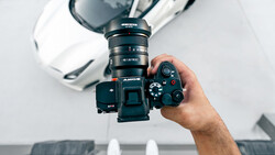 Photographer holding a mirrorless camera with mounted lens and external flash against a white backdrop.