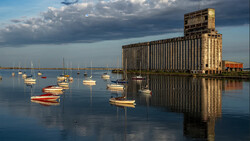 Sailboats moored in calm water with a large industrial grain elevator reflected in the glassy surface at golden hour.