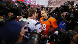 Football player in orange jersey surrounded by photographers and media at stadium field level.