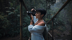 Woman in light blue off-shoulder dress holding vintage film camera in lush forest setting.