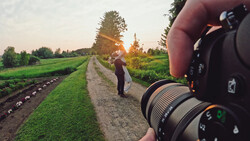 Photographer holding camera with telephoto lens, photographing person standing on rural dirt road at golden hour.