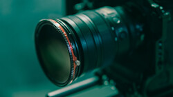 Close-up of a telephoto lens with red focus ring mounted on a camera body against a teal-colored background.