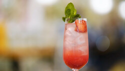 Red cocktail with ice, mint garnish, and strawberry slice in tall glass.