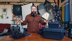 Bearded man in burgundy shirt standing at wooden desk surrounded by photography lighting equipment and cases.