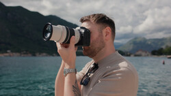 Photographer with telephoto lens photographing across a lake toward mountains on a partly cloudy day.