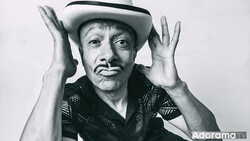 Man in cowboy hat and patterned shirt with hands raised to sides of head, black and white portrait.