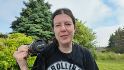 Photographer holding a vintage SLR camera with manual lens, smiling outdoors with trees and green fields in background.