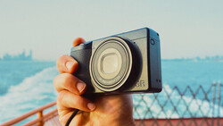 Hand holding compact digital camera with extended zoom lens, photographing coastal landscape with water and distant shoreline.