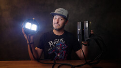 Photographer holding a portable LED light panel in one hand and lighting equipment in the other against a dark background.