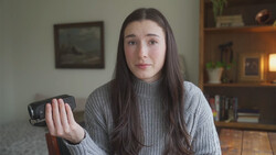 Young woman in gray ribbed sweater holding small handheld device toward camera in home interior.