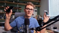 Young content creator holding multiple smartphones while speaking into a microphone at a desk setup.