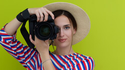 Woman in striped shirt and straw hat holding DSLR camera up to her face against yellow background.