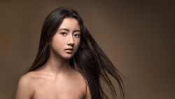 Woman with long black hair against neutral brown background, looking toward camera.