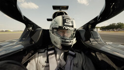 Driver in racing helmet and suit inside open-cockpit race car, captured from above on runway.