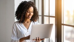 Woman with curly dark hair wearing a light gray button-up shirt, holding and looking at an open laptop while standing near a sunlit window.