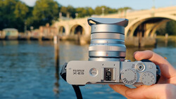 Fujifilm X-E5 camera with silver lens held in hand over a river and stone bridge.