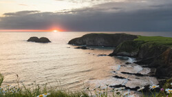 Sunset over a rugged coastal landscape with green cliffs, rocky outcrops, and small islands.