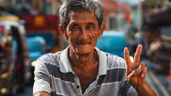 Elderly man with gray hair smiling and making a peace sign gesture at camera.