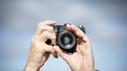Photographer holding a rangefinder camera up to eye level against a cloudy sky backdrop.