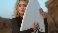 Blonde woman in black wetsuit holding white surfboard at rocky coastline.