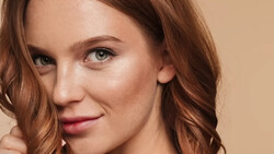 Woman with wavy auburn hair and green eyes against a warm beige background.