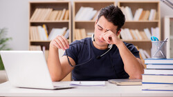 Man in dark shirt sitting at desk with laptop, holding glasses and looking thoughtfully at papers.