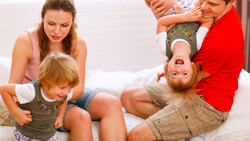 Family of four playing together on a white bed, with parents and children laughing and playfully interacting.