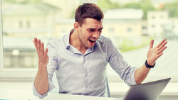 Man at desk smiling while looking at laptop screen with hands raised expressively.