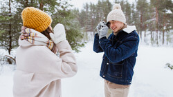 Two people playing in snow, one in a white jacket and yellow beanie, the other in a denim jacket and white beanie, with evergreen trees and snowy landscape in background.