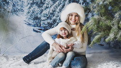 Woman and child sitting together in snow wearing matching cream-colored winter hats and sweaters.