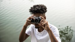 Person holding vintage rangefinder camera up to face, positioned by water with natural overcast lighting.