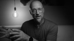 Man with glasses wearing a dark t-shirt gestures while speaking, lit by a hanging incandescent bulb against a dark background.