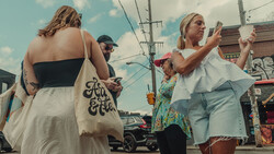 Woman holding a canvas tote bag with script lettering walks down a sunny street with other pedestrians.