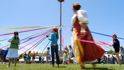 Children and adults holding colorful ribbons attached to a tall maypole during an outdoor spring celebration on a sunny day.