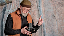 Older man with beard and glasses holding a vintage film camera against a textured stone wall.