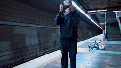Man in dark suit and sunglasses holding camera at subway station platform.