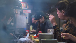Woman holding chopsticks over a bowl in a smoky kitchen environment with other diners visible in background.