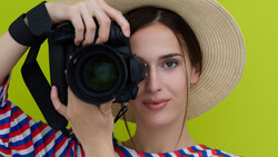 Photographer wearing a striped shirt and straw hat holds a DSLR camera up to her eye against a bright yellow-green background.