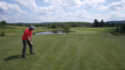 Golfer in red shirt taking a shot on a wide-open fairway with a pond and tree line visible in the distance.