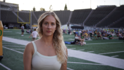 Portrait of a woman with blonde wavy hair on a college football field during an event with crowds in the stadium background.