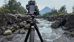 Professional camera mounted on tripod overlooking a rushing mountain river with volcanic peak in background.