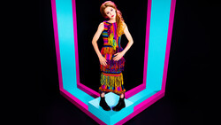 Woman in colorful fringed outfit posed against bright cyan and magenta geometric neon lines.