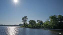 Bright sunlit river landscape with trees lining the far bank under clear blue sky.