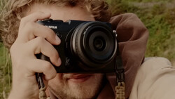 Photographer holding a Fujifilm mirrorless camera up to his face to compose a shot outdoors.
