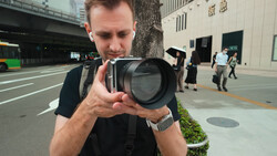 Photographer holding a large telephoto lens while looking through the viewfinder on an urban street.