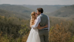 Couple in wedding attire kissing on a hillside overlooking forested valley.