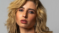 Portrait of a woman with shoulder-length wavy blonde hair against a neutral gray background.