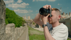 Male photographer holding a compact camera to his eye, composing a shot along a stone fortification wall.