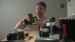 Photographer examining a vintage film camera while surrounded by other classic cameras on a wooden desk.