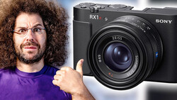 Man in purple shirt with curly hair and glasses gesturing toward a Sony RX1 II compact camera.
